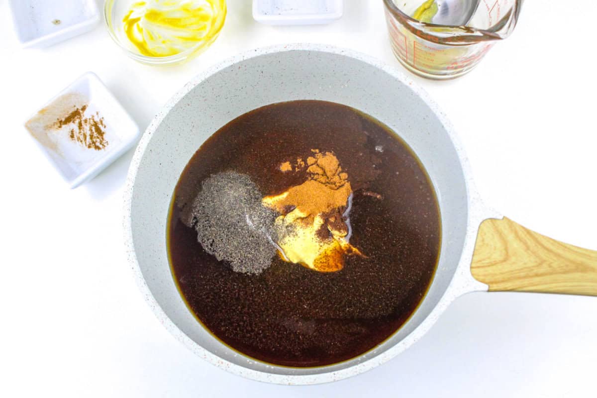 A white saucepan with a wooden handle contains a dark brown liquid for bourbon maple glazed ham, with spices sprinkled on top. Nearby are small bowls with various ingredients and a glass measuring cup.