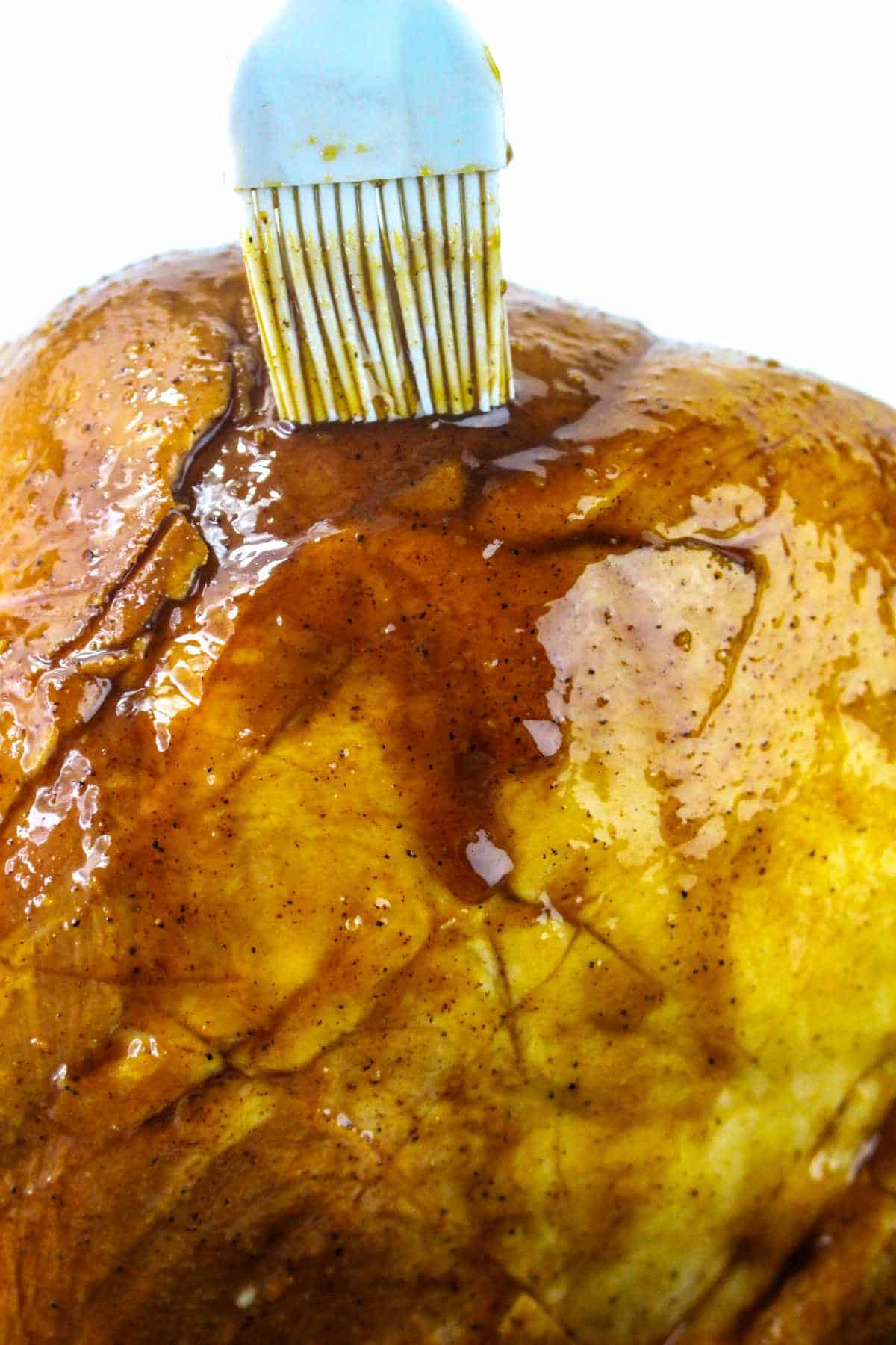 A close-up of a ham surface being brushed with a shiny, brown glaze, reminiscent of the rich coating on a maple glazed ham, using a white pastry brush.