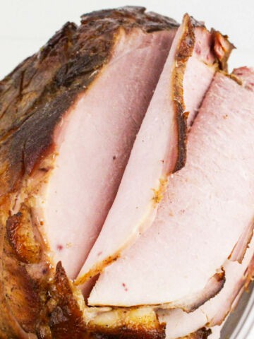 A close-up of a bourbon maple glazed ham with several thick slices cut, showing the juicy, pink interior and browned, crispy edges. The ham is on a white plate, garnished with green herbs.