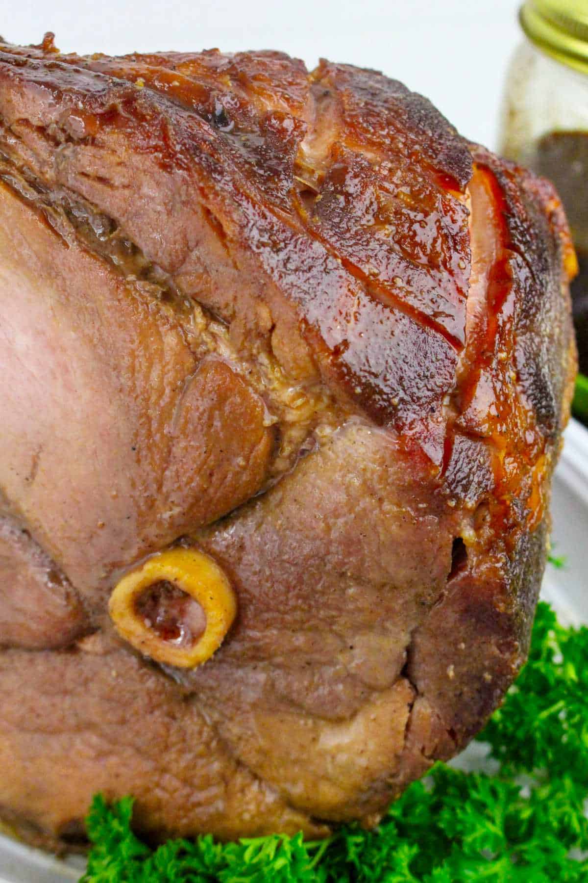 Close-up of a bourbon maple glazed ham with a visible bone, sitting on a white plate garnished with fresh parsley.