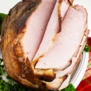 A bourbon maple glazed ham, spiral-sliced and garnished with fresh parsley, sits on a white plate atop a red and beige plaid cloth.