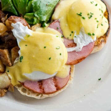 Close-up of eggs Benedict with hollandaise sauce: two poached eggs on Canadian bacon and English muffins, topped with fresh chives, served alongside potatoes and greens—perfect inspiration for breakfast recipes.