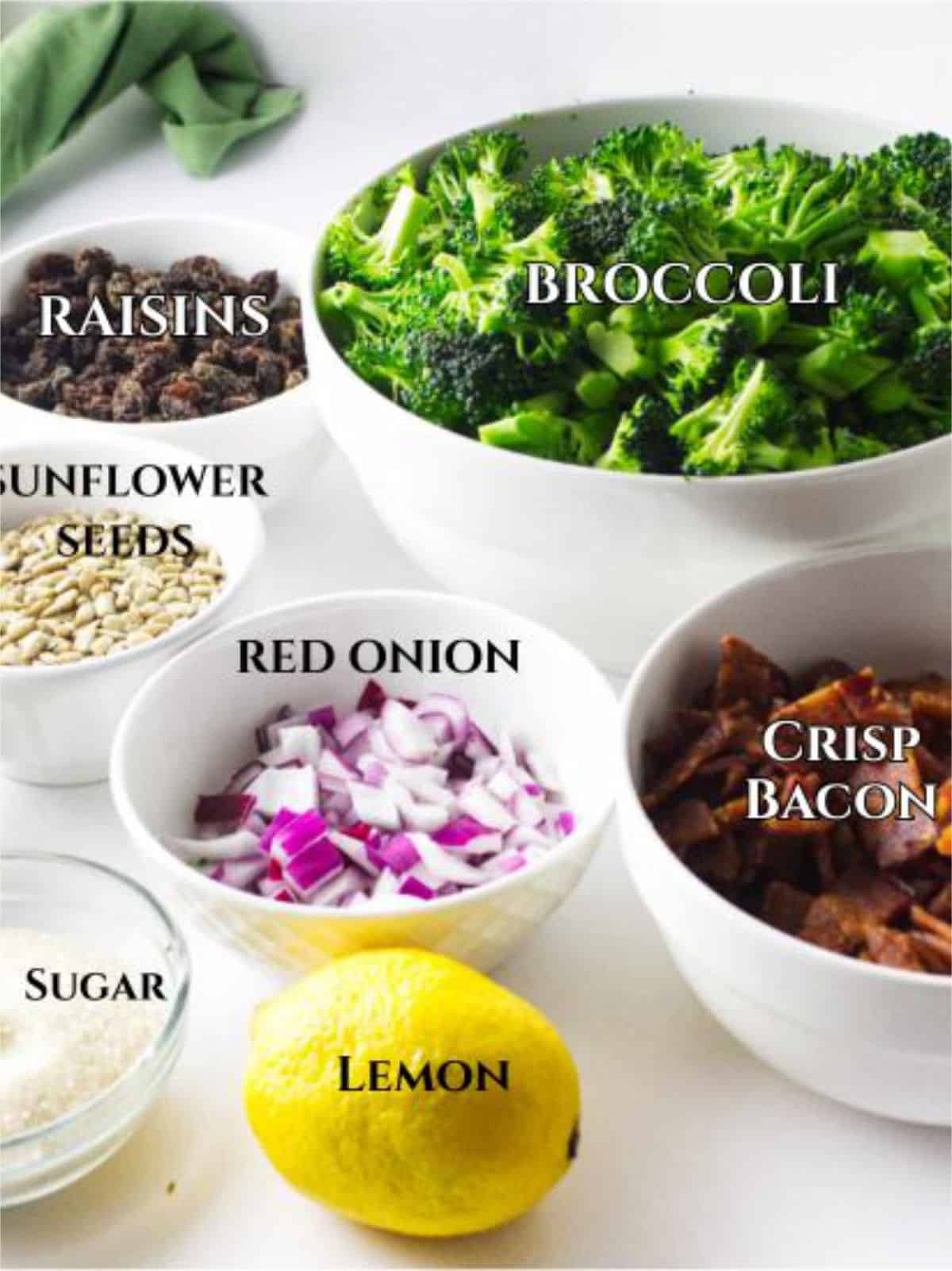 A display of ingredients for a classic Walmart broccoli salad in bowls, labeled: broccoli, raisins, sunflower seeds, chopped red onion, crisp bacon, sugar, and a whole lemon.