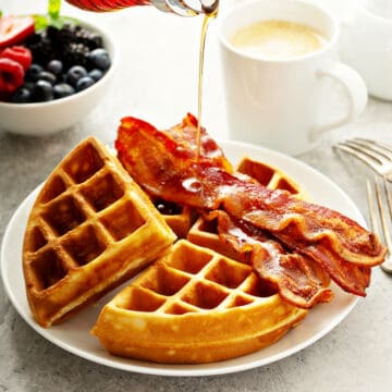 A plate of golden waffles topped with crispy bacon is being drizzled with syrup. Perfect for brunch recipes, the scene includes a cup of coffee and a bowl of fresh berries in the background, with forks beside the plate.