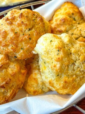 Golden, fluffy cheddar biscuits with flecks of herbs are piled in a white cloth-lined basket.