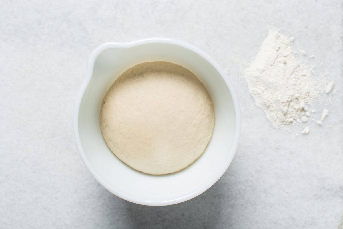 A white bowl with risen bread dough, perfect for making Chinese steamed buns, sits on a light surface, with a small pile of flour nearby.