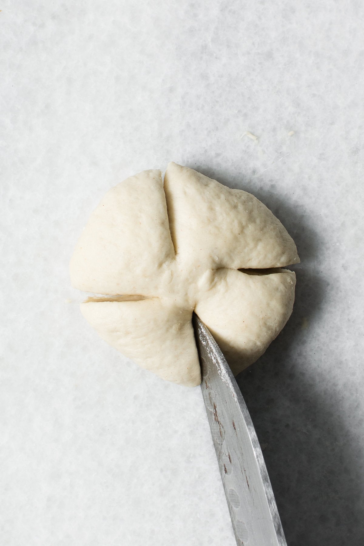 A knife making four evenly spaced cuts on a round ball of dough for chinese steamed buns, placed on a white surface.