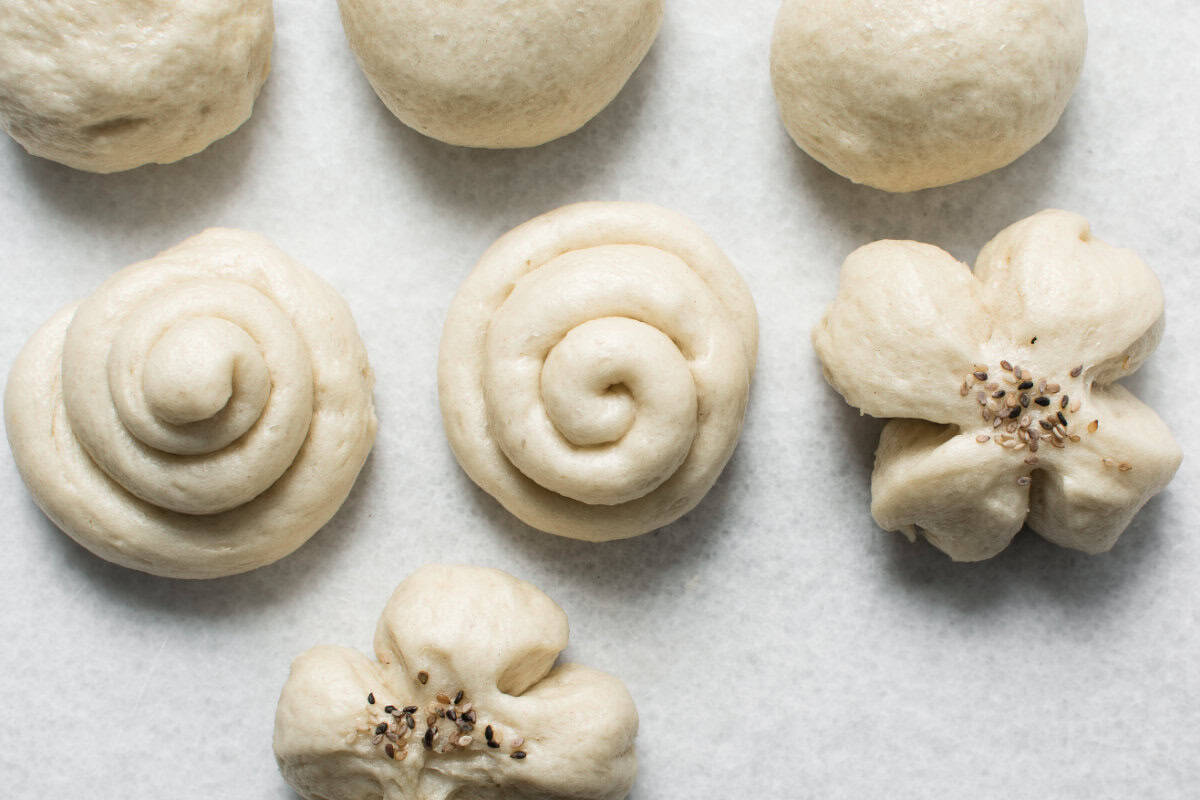 Six pieces of unbaked dough, resembling balls, spirals, and flower-like forms, are arranged on a white parchment-lined surface. Two dough pieces, similar to Chinese steamed buns, are decorated with seeds in the center.