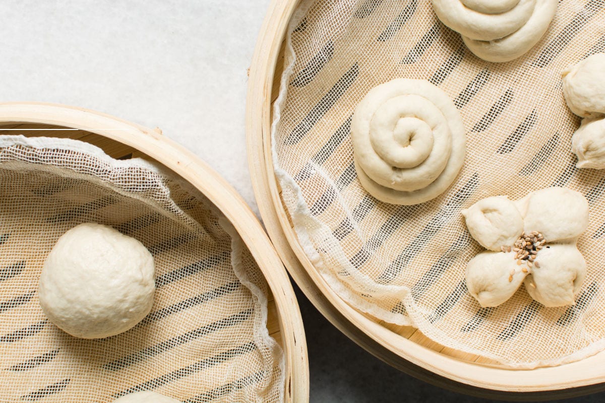 Two round bamboo steamers lined with mesh hold unsteamed dough shapes—like a ball, a spiral, and a flower—that will soon become delicious Chinese steamed buns on a light surface.