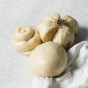 Three Chinese steamed buns of different shapes rest on a light surface-one round, one twisted like a rose, and one flower-shaped with seeds on top; a white cloth is partially visible in the corner.