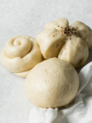 Three Chinese steamed buns of different shapes rest on a light surface-one round, one twisted like a rose, and one flower-shaped with seeds on top; a white cloth is partially visible in the corner.
