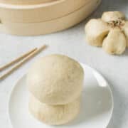 Two Chinese steamed buns stacked on a white plate, with wooden chopsticks beside them. In the background, a bamboo steamer holds more Chinese steamed buns and a flower-shaped bun on a light surface.