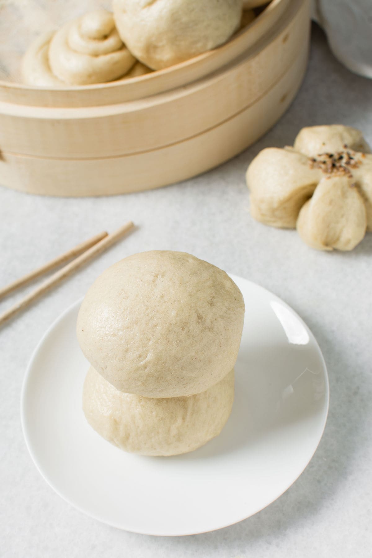 Two Chinese steamed buns stacked on a white plate, with wooden chopsticks beside them. In the background, a bamboo steamer holds more Chinese steamed buns and a flower-shaped bun on a light surface.