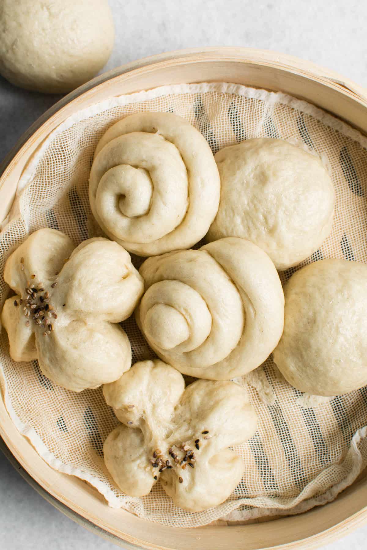 A bamboo steamer lined with cloth holds several chinese steamed buns in different shapes, including round, spiral, and flower-like forms. The chinese steamed buns appear soft and freshly cooked.