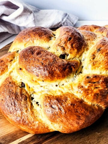 Golden-brown, braided chocolate brioche loaf sits on a wooden cutting board, with a towel in the background.