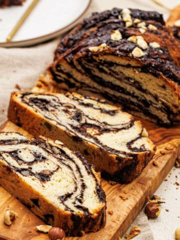 Sliced chocolate hazelnut babka with visible chocolate swirls, served on a wooden cutting board and garnished with chopped nuts.