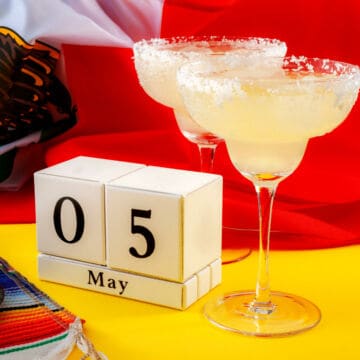 Two margarita glasses with salted rims sit beside a calendar displaying "05 May," with a colorful cloth and a red, white, and green flag in the background—perfect inspiration for trying new Cinco de Mayo recipes.