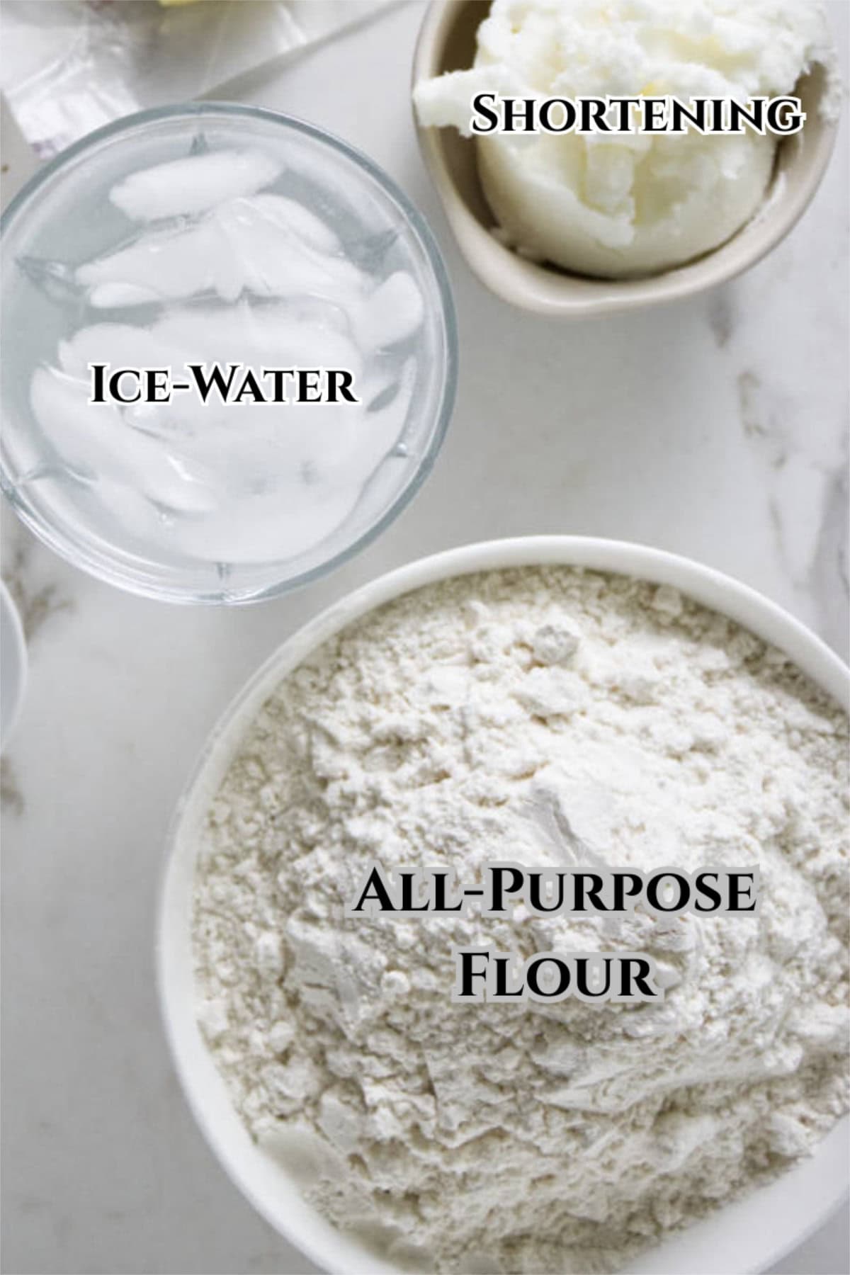 A bowl of all-purpose flour, a bowl of shortening, and a glass of ice water are arranged on a white surface-just like the classic Marie Callender pie crust recipe. Each item is labeled with its name in bold text.