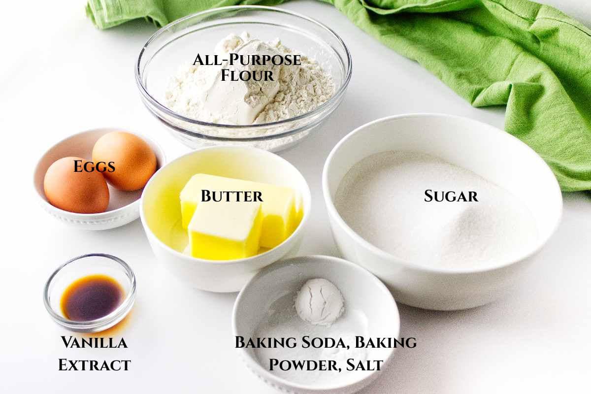Baking ingredients arranged in bowls: all-purpose flour, sugar, butter, eggs, baking soda, baking powder, salt, and vanilla extract, labeled with text. Green cloth in the background.