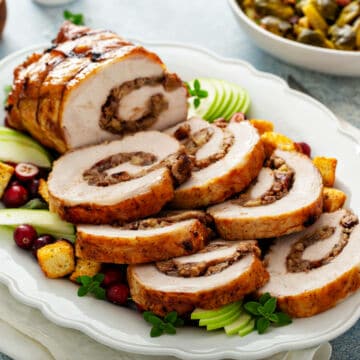 Sliced stuffed turkey roulade arranged on a white platter with fresh herbs, apple slices, cranberries, and croutons—perfect for those seeking impressive dinner recipes—with a bowl of vegetables in the background.