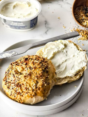 A sliced everything bagel on a white plate, with one half spread with cream cheese. A knife with cream cheese rests on the plate, and a container of cream cheese and a bowl of everything bagels recipe seasoning are in the background.