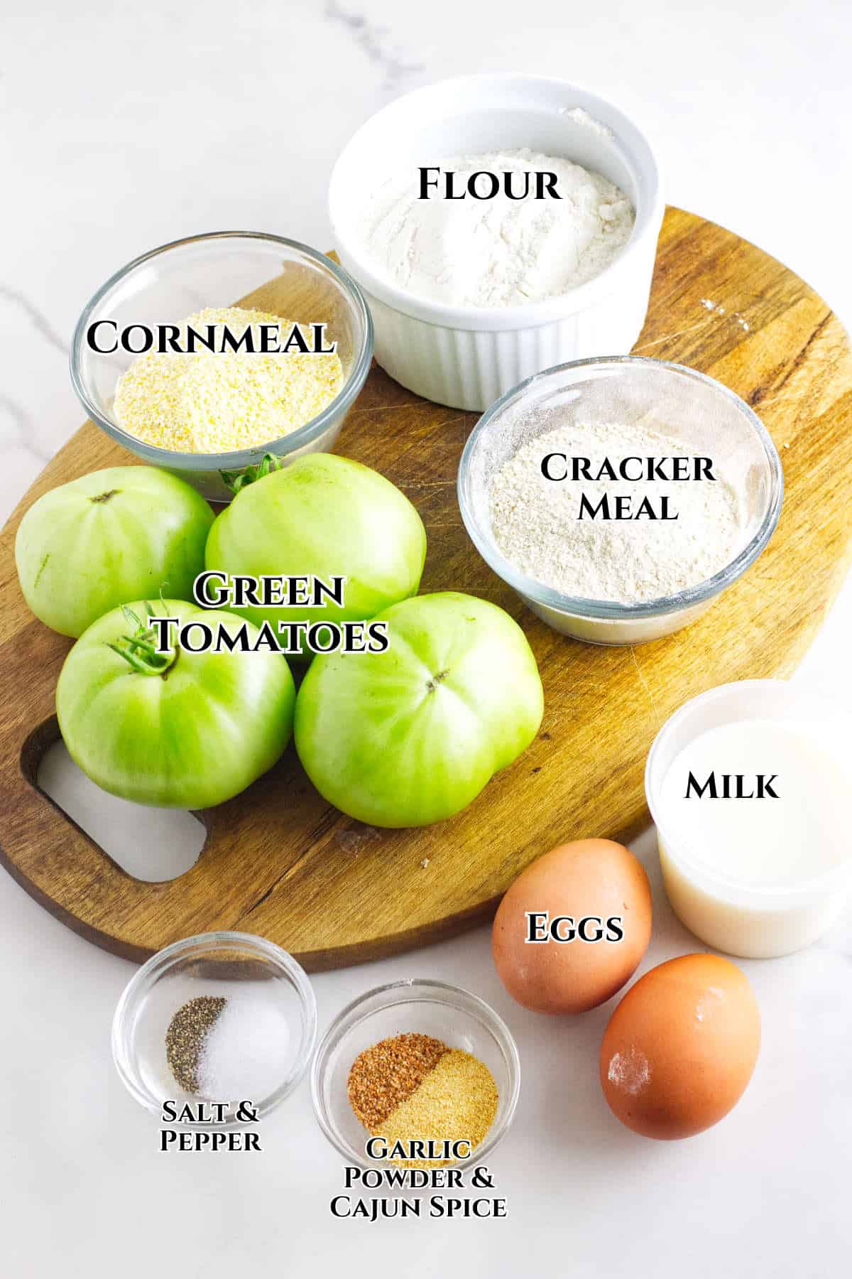 A wooden board displays ingredients for a fried green tomatoes recipe: green tomatoes, bowls of flour, cornmeal, cracker meal, milk, and two eggs. Small labeled bowls hold salt & pepper and garlic powder & Cajun spice.