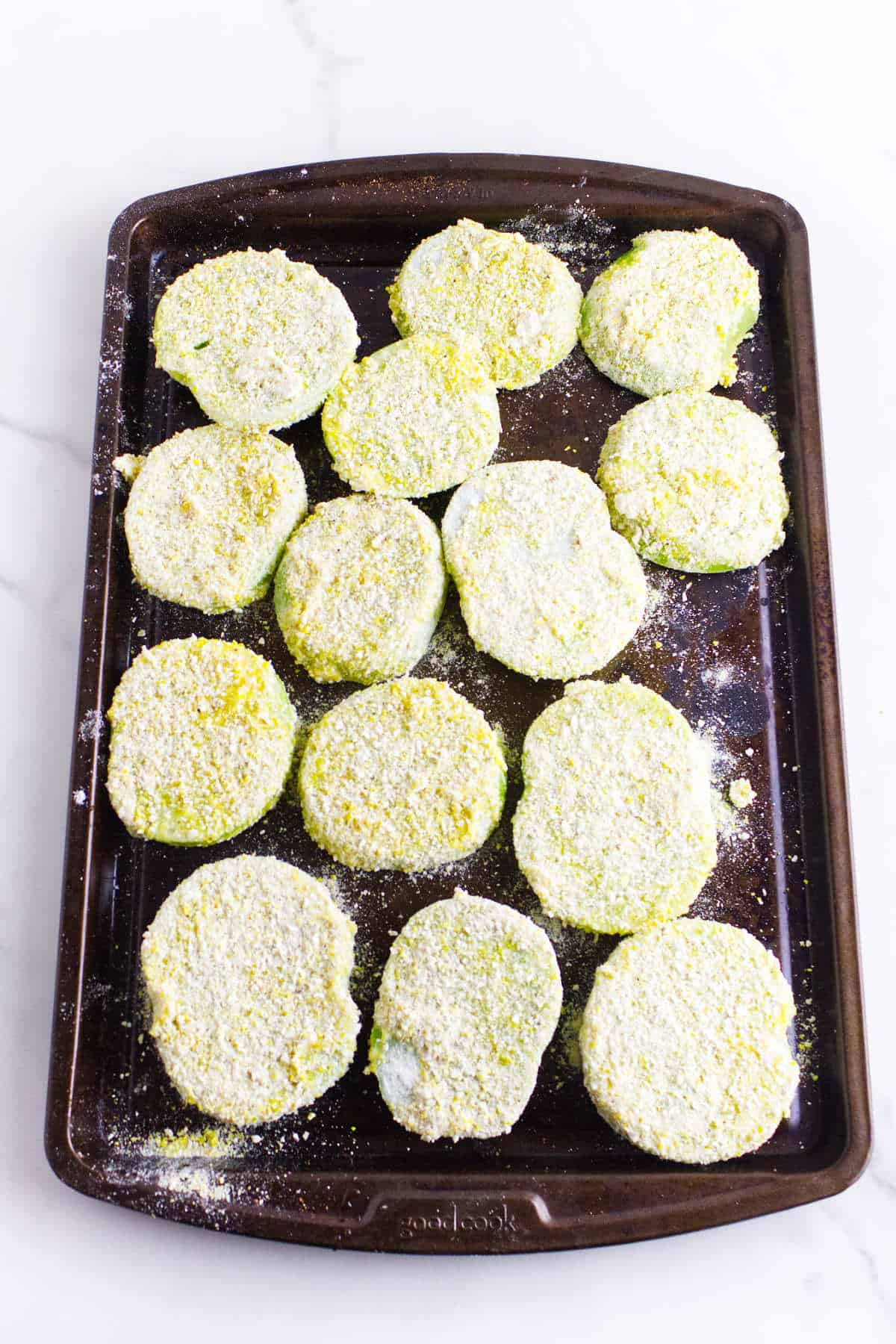 A baking tray with several breaded, uncooked slices evenly spaced and coated in a yellow cornmeal mixture, ready to be baked or fried.