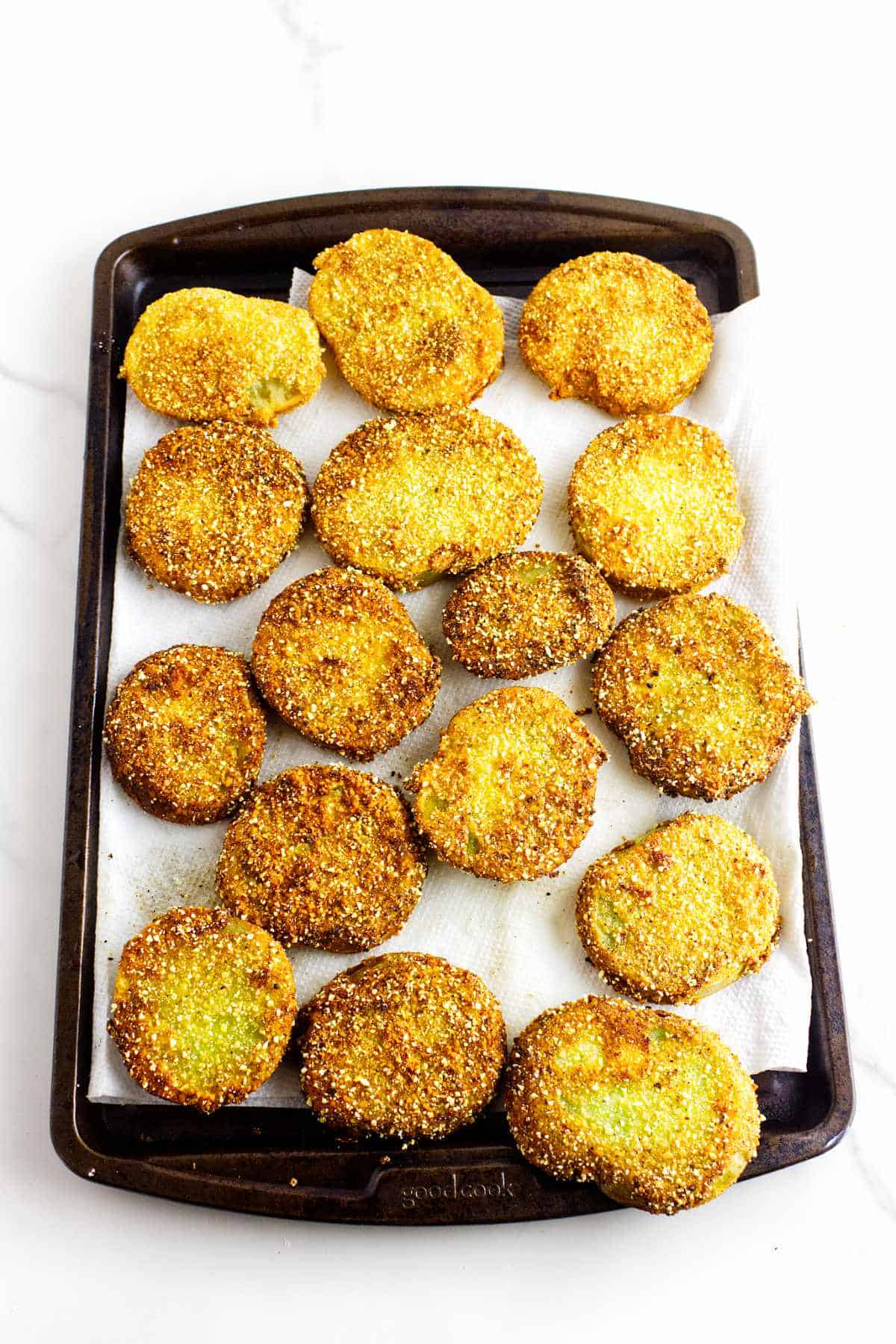 A baking tray lined with paper towels holds freshly fried slices of tomato coated in a golden.