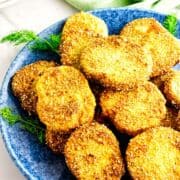 A blue plate filled with crispy, golden-brown fried green tomatoes, garnished with sprigs of fresh dill, sits on a white surface near a green cloth and stacked white plates in the background.