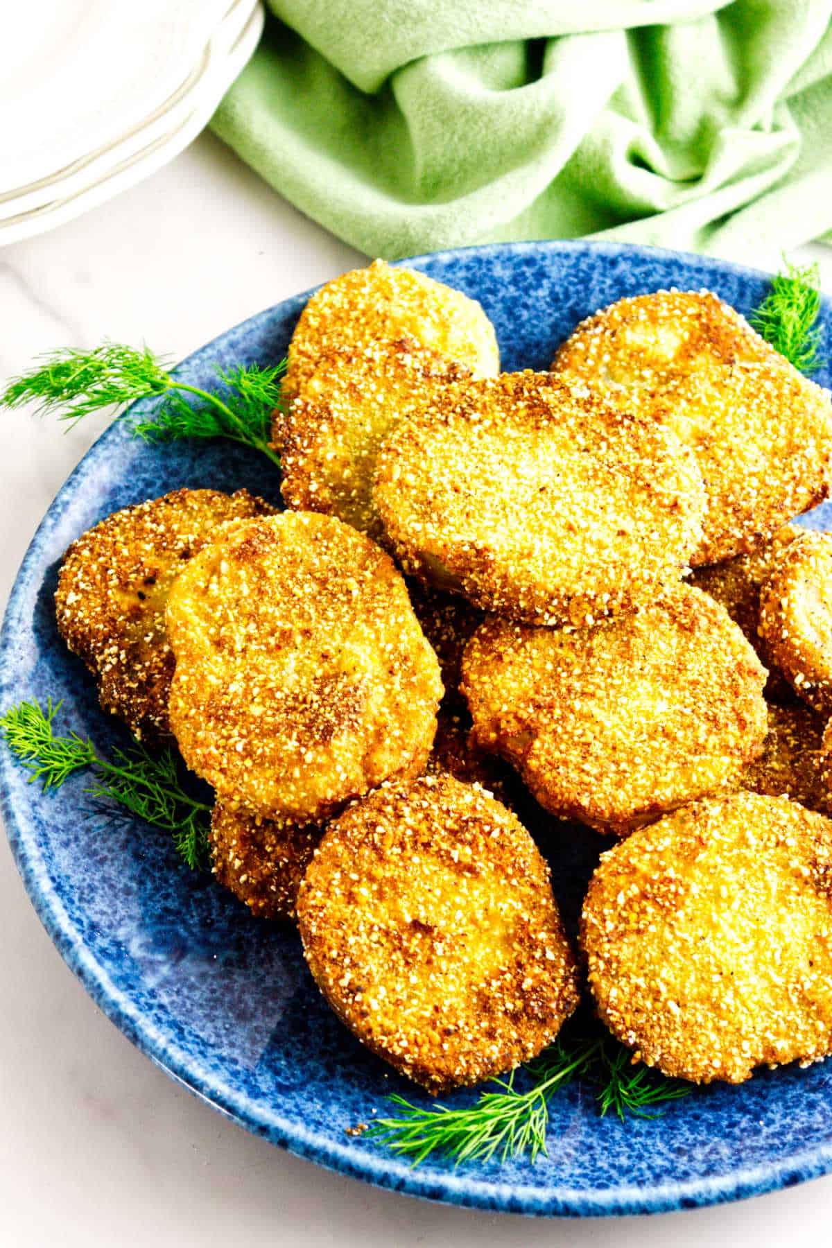 A blue plate filled with crispy, golden-brown fried green tomatoes, garnished with sprigs of fresh dill, sits on a white surface near a green cloth and stacked white plates in the background.
