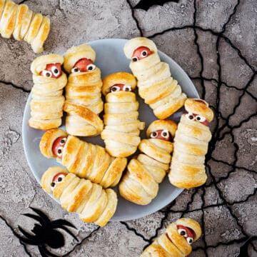 A plate of mini mummy hot dogs, a fun take on Halloween recipes, features hot dogs wrapped in puff pastry with edible eyes. The spooky snacks are served on a cracked concrete surface decorated with black spiders.