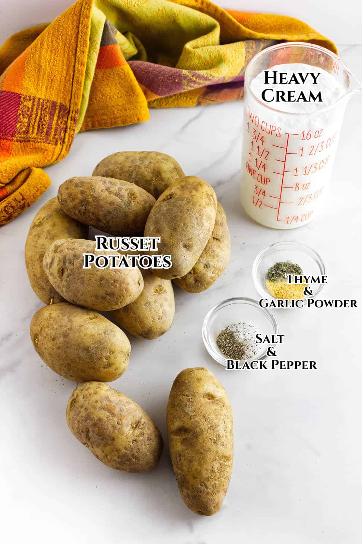 Several russet potatoes are arranged on a white surface with small bowls of thyme, garlic powder, salt, and black pepper nearby—perfect for making one bowl scalloped potatoes. A measuring cup of heavy cream and a colorful kitchen towel add brightness to the scene.