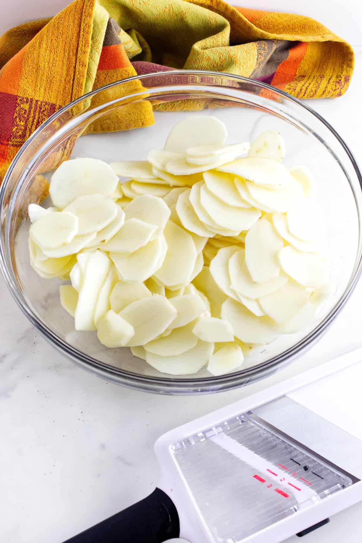 A glass bowl filled with thinly sliced potatoes sits on a white surface next to a white mandoline slicer, with a colorful orange and yellow kitchen towel in the background.