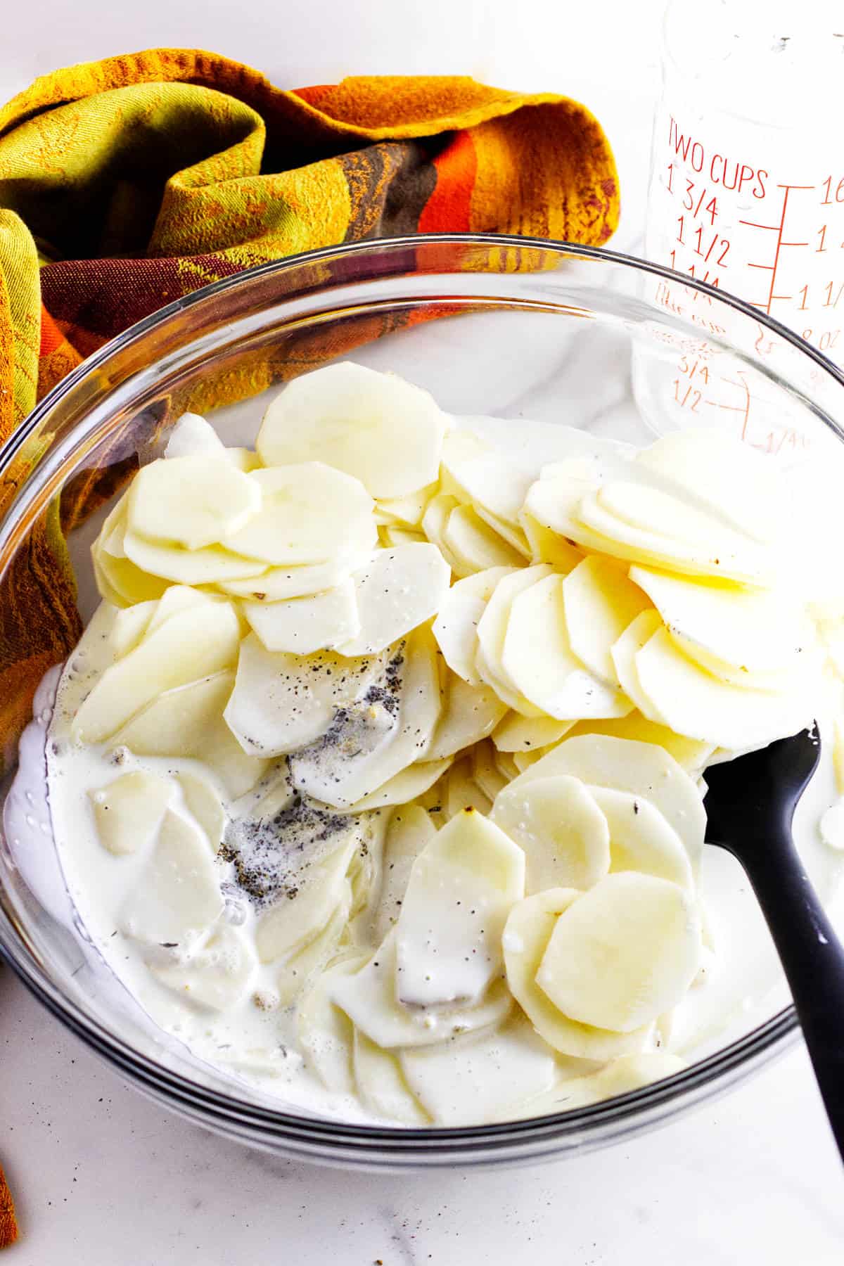 A glass bowl filled with sliced potatoes mixed with cream and pepper, a black spoon, an orange and yellow cloth, and a clear measuring cup are on a white surface.