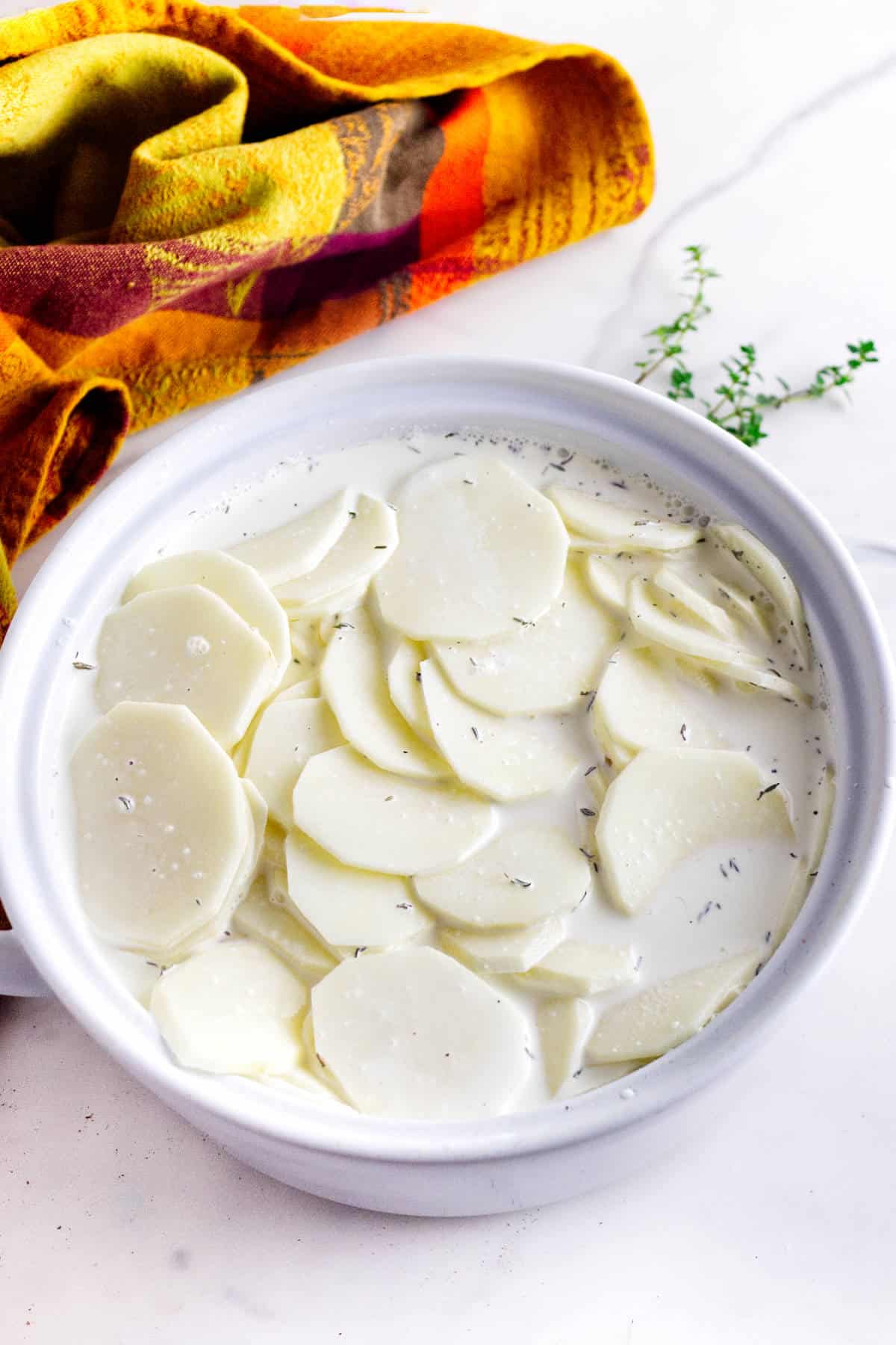 Sliced potatoes soaking in a creamy white liquid with herbs, in a white bowl on a marble surface. A colorful yellow and orange cloth and a sprig of thyme are nearby.