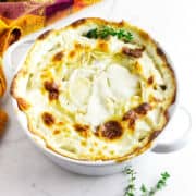 A white baking dish filled with creamy, golden-brown scalloped potatoes, garnished with fresh thyme.