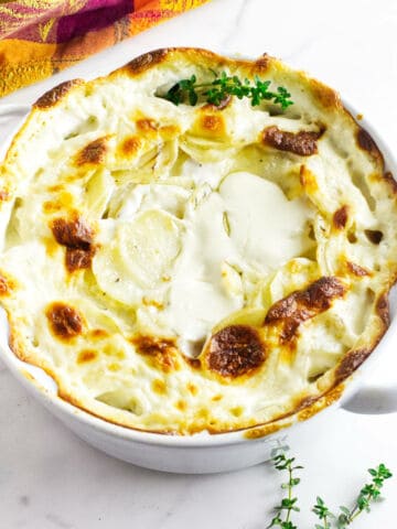 A white baking dish filled with creamy, golden-brown scalloped potatoes, garnished with fresh thyme.