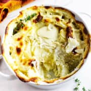 A baked potato gratin in a white dish, with a golden-brown cheesy top and a portion scooped out, revealing creamy potato layers. Fresh thyme sprigs are placed on the dish and the marble surface nearby. A colorful towel is in the background.
