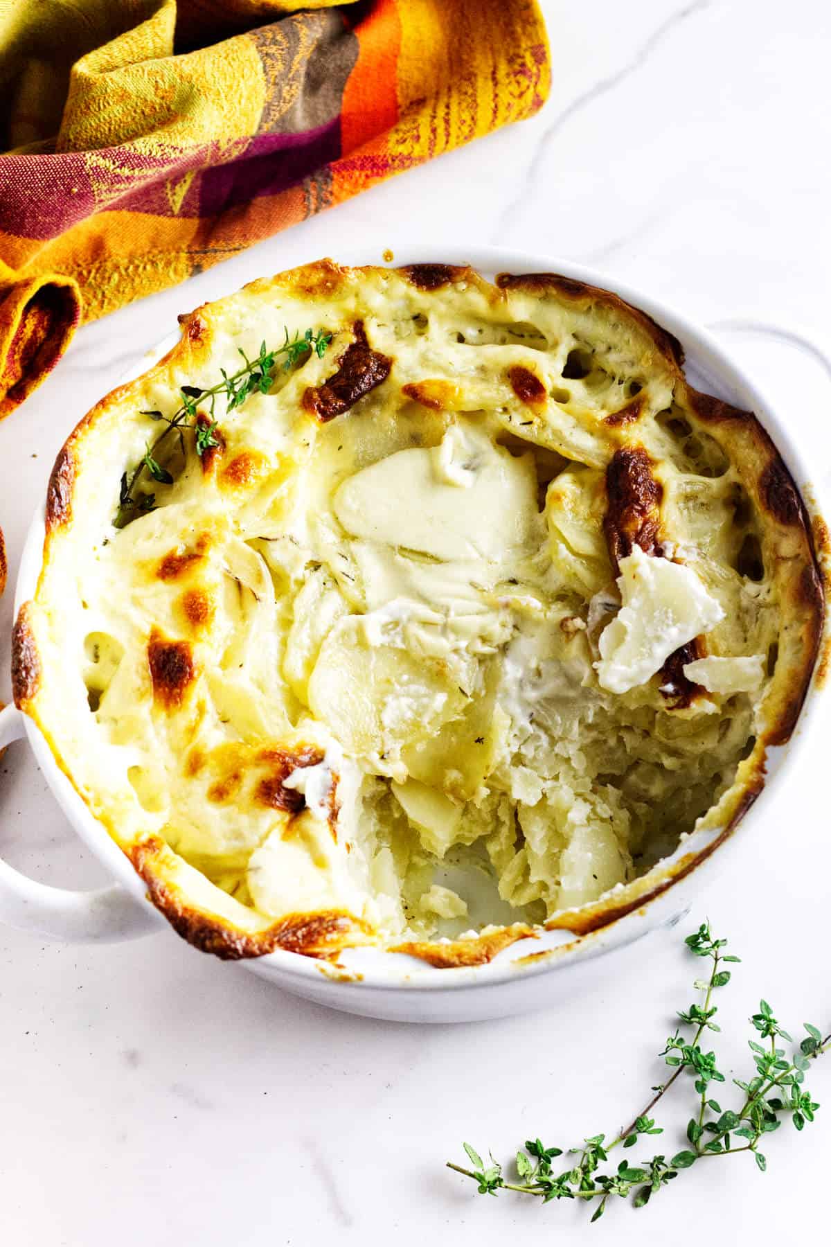 A baked potato gratin in a white dish, with a golden-brown cheesy top and a portion scooped out, revealing creamy potato layers. Fresh thyme sprigs are placed on the dish and the marble surface nearby. A colorful towel is in the background.