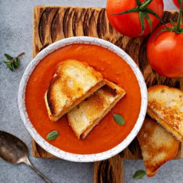 A bowl of creamy tomato soup topped with two slices of toasted bread sits on a wooden board next to fresh tomatoes and a tarnished spoon. Small basil leaves garnish the soup, making it a perfect pick for classic lunch recipes.