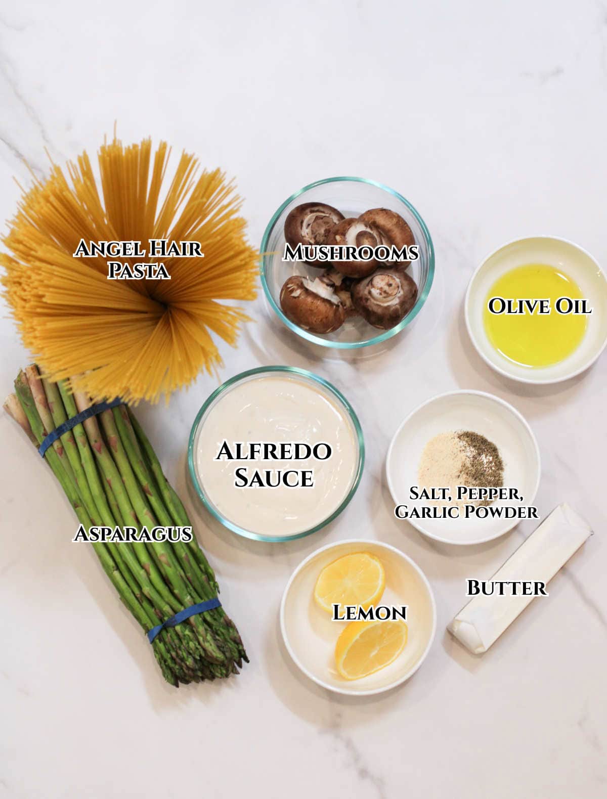 Top view of ingredients for mushroom asparagus pasta on a marble surface: uncooked angel hair, mushrooms, asparagus, olive oil, Alfredo sauce, lemon slices, salt, pepper, garlic powder, and a pat of butter—all clearly labeled.