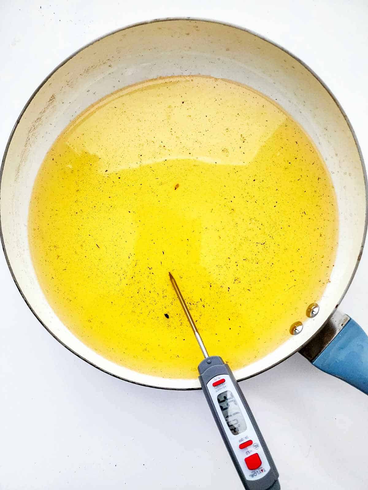 A frying pan filled with hot oil, perfect for making New Orleans beignets, with a digital thermometer inserted to check the temperature. The oil appears clear with some small black specks, and the pan has a blue handle.