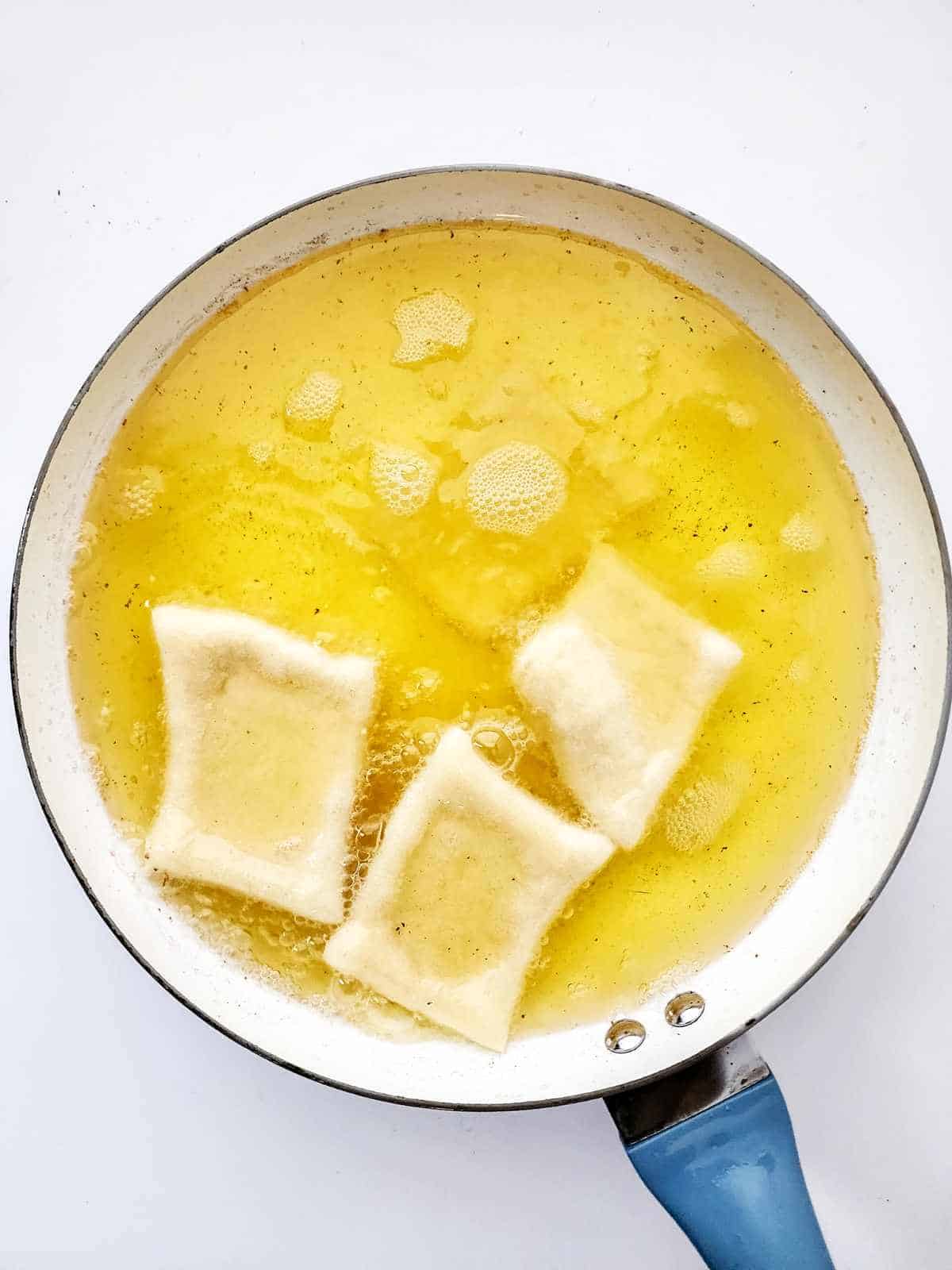 A frying pan with hot oil contains three pieces of ravioli frying, surrounded by bubbles like New Orleans beignets, against a white background.
