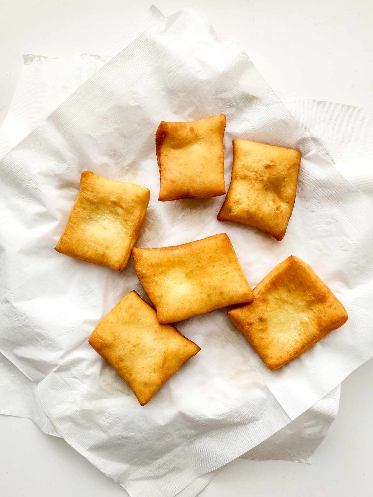 Six golden-brown New Orleans beignets rest on a white paper towel, arranged casually on a flat surface. The dough squares look crisp and slightly puffed.