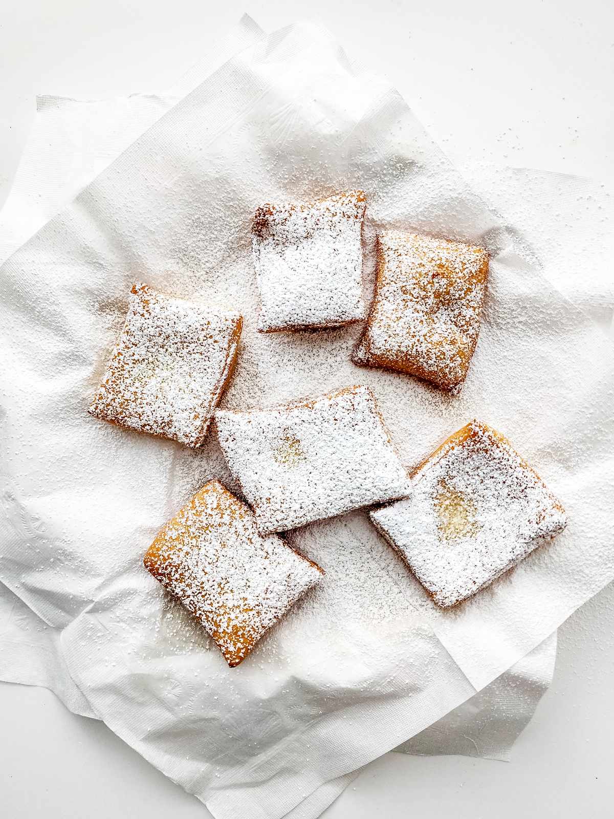 Six square New Orleans beignets dusted with powdered sugar sit on a piece of white parchment paper against a white background.