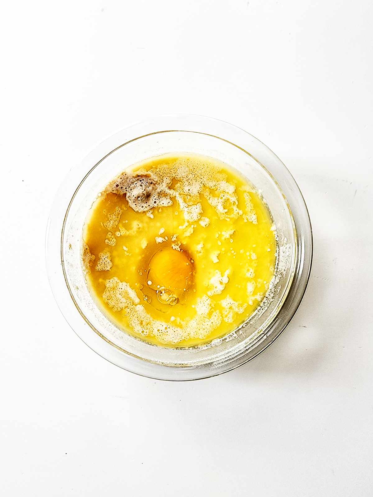 A clear glass bowl on a white surface contains a cracked raw egg, melted butter, and brown sugar-ingredients often used in classic New Orleans beignets-ready to be mixed.