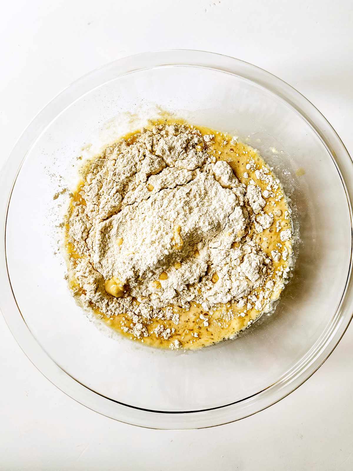 A glass mixing bowl contains a partially mixed combination of flour and a yellowish wet batter, ready to be transformed into classic New Orleans beignets, on a white surface.