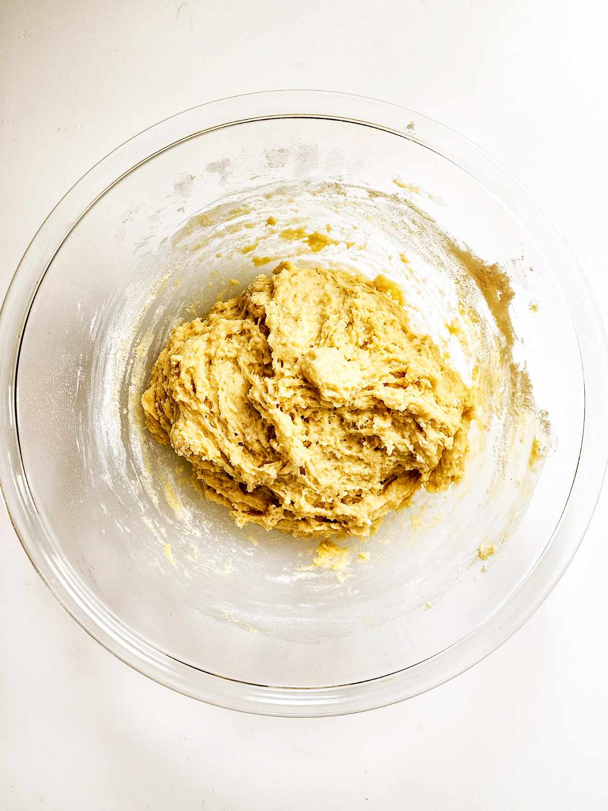 A clear glass bowl on a white surface contains a thick, partially mixed New Orleans beignets dough, with visible flour around the edges inside the bowl.