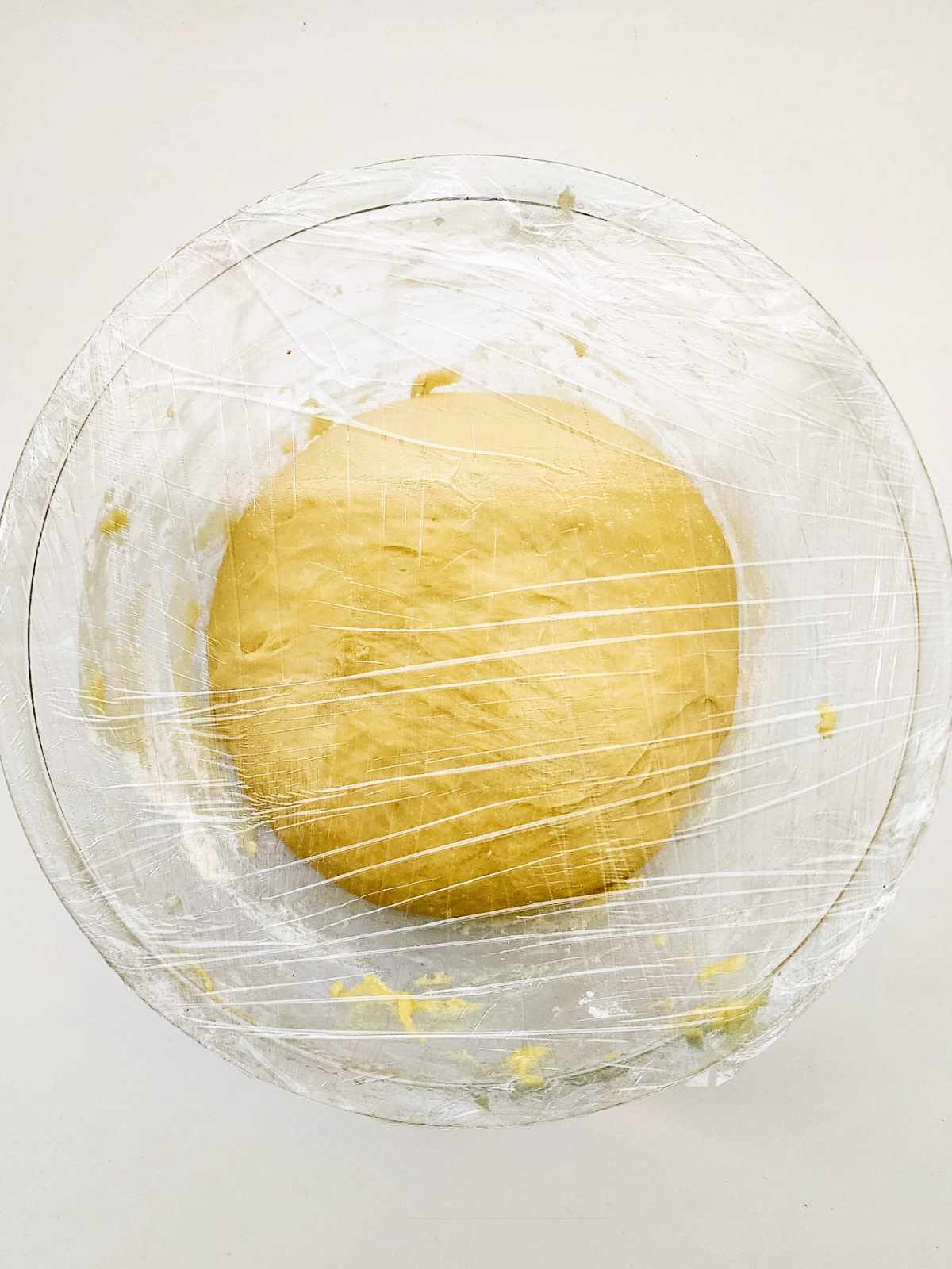 A glass bowl with dough inside, covered with plastic wrap. The smooth dough is rising, ready to become classic New Orleans beignets, set against a plain white background.