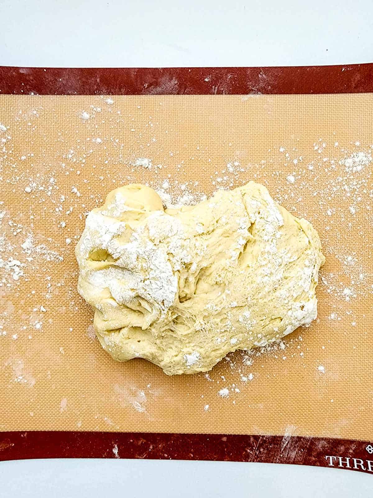 A mound of raw, lightly floured New Orleans beignets dough rests on a silicone baking mat atop a white surface, with scattered flour around it.